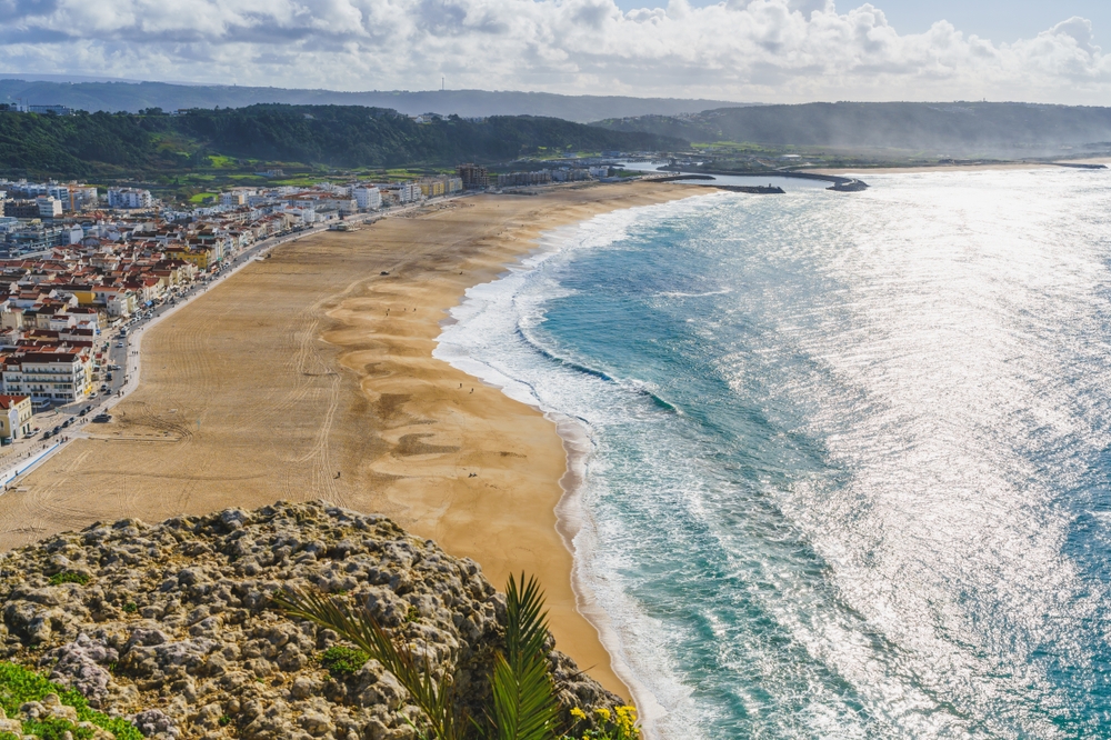 Widok na plażę, ocean i miasto Nazare z wysokiego punktu widokowego ze słońcem lśniącym na wodzie. Srebrne Wybrzeże, Costa de Prata, Portugalia

